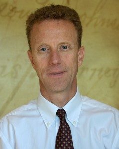 Closeup of a smiling middle aged white man with short-cropped light brown hair, wearing a navy suit, white shirt, and navy tie with small white polka-dots