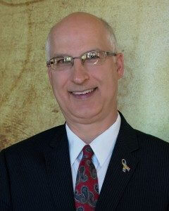 Dave Ballou from the chest up smiling wearing a black suit with a red and blue tie.