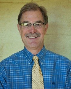 A portrait of Lee Romano from the chest up. He is smiling and wearign a blue plaid button up shirt with a yellow tie.