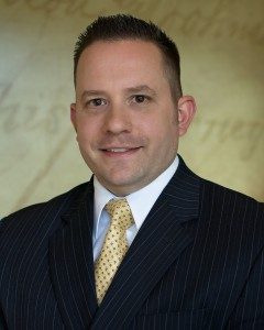 Nathanial Marcoux from the chest up smiling while wearing a black suit with a yellow-golden tie.