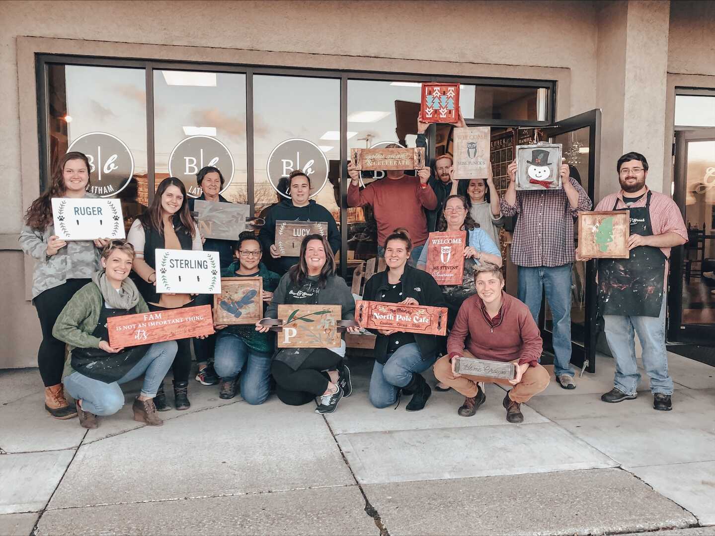 A group of people hold up custom wood sign they all made in a workshop.