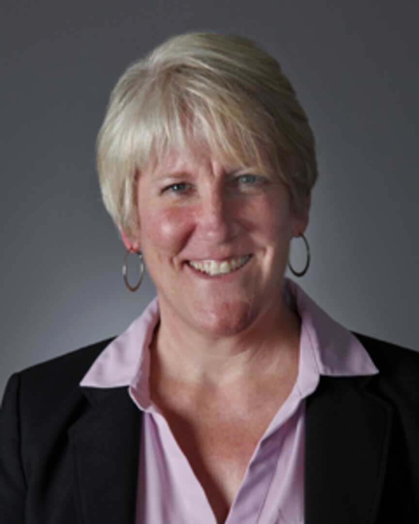 Closeup of a smiling middle aged white woman with short blond hair, wearing hoop earrings, a light blue open-collared blouse, and navy jacket