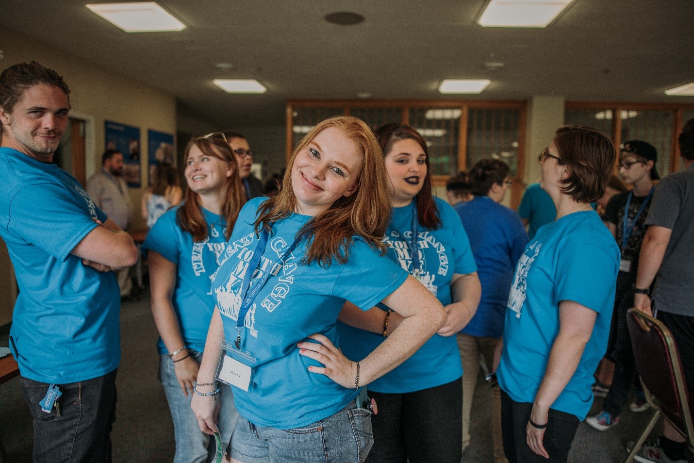 A teenage girl with red hair smiles for the camera surrounded by other kids her age. They are all wearing light blue shirts and are socializing among themselves at an event.
