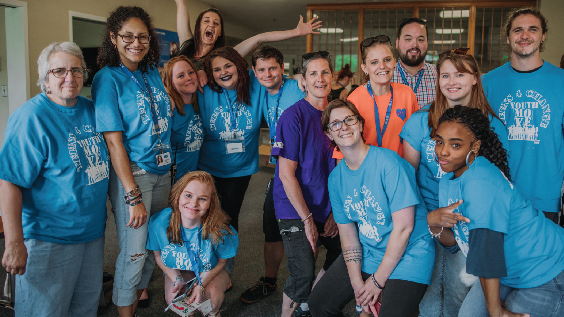 A group of 14 teens and adults wearing matching light blue t-shirts. Some are jumping, throwing up peace signs, laughing, and crouching to get in the frame.