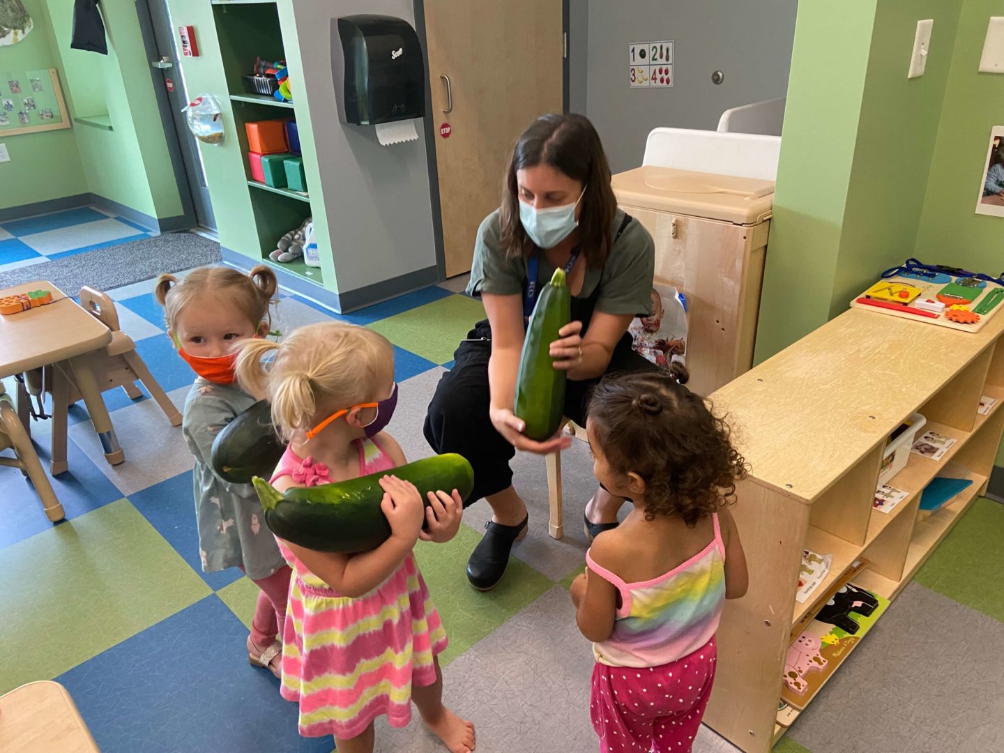 A teacher shows three students a large zucchini that they grew in their community garden. The zucchinis can barely be held by the kids!