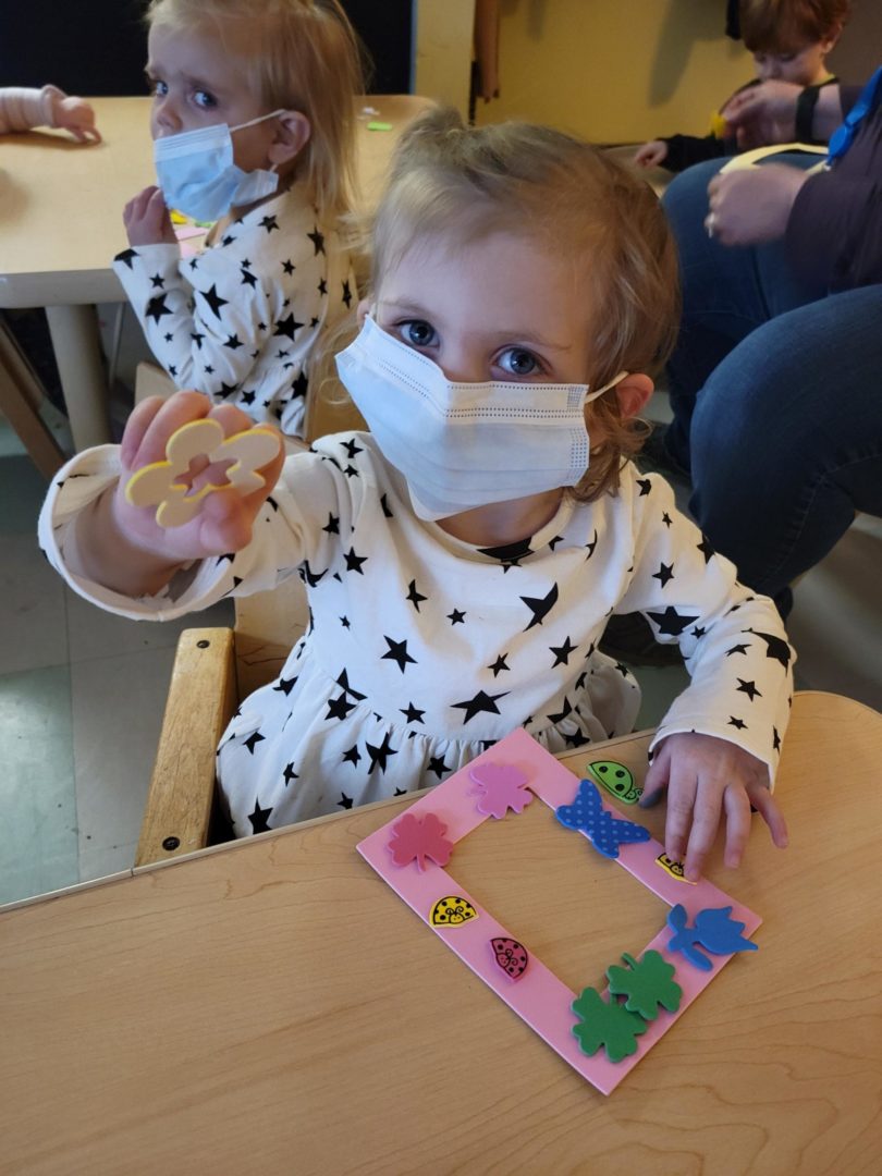 Young child wearing a mask holding up artwork. The artwork looks like a yellow foam flower that can be glued to paper alongside other foam shapes of varying colors.