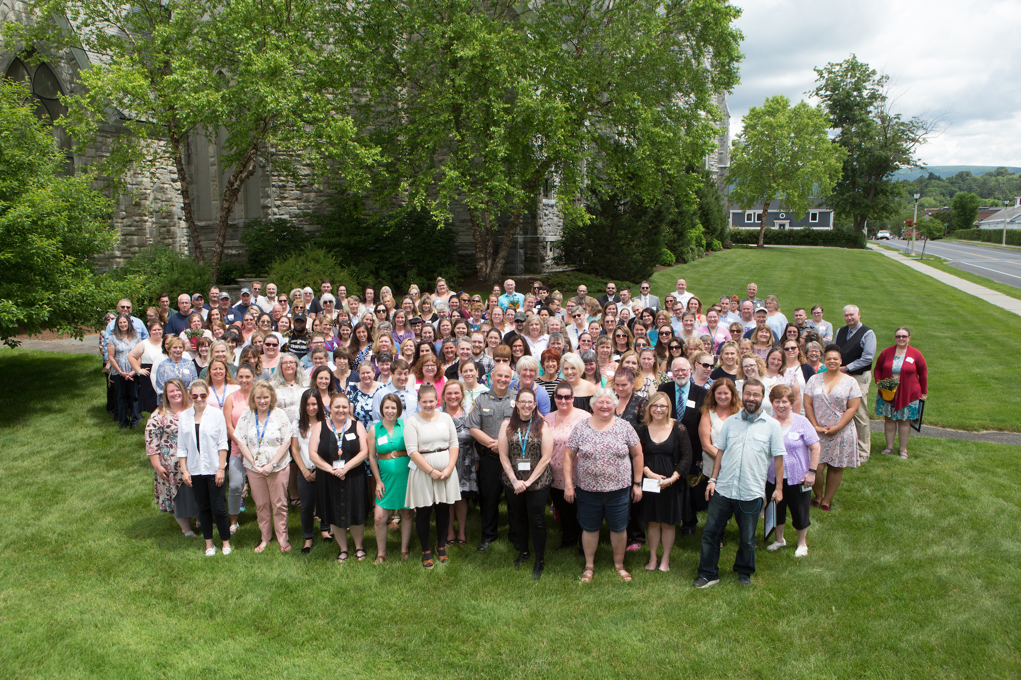 A very large group of people pose for a picture on a green lawn. There are trees in the background and Main Street in Bennington, Vermont can be seen off to the side.