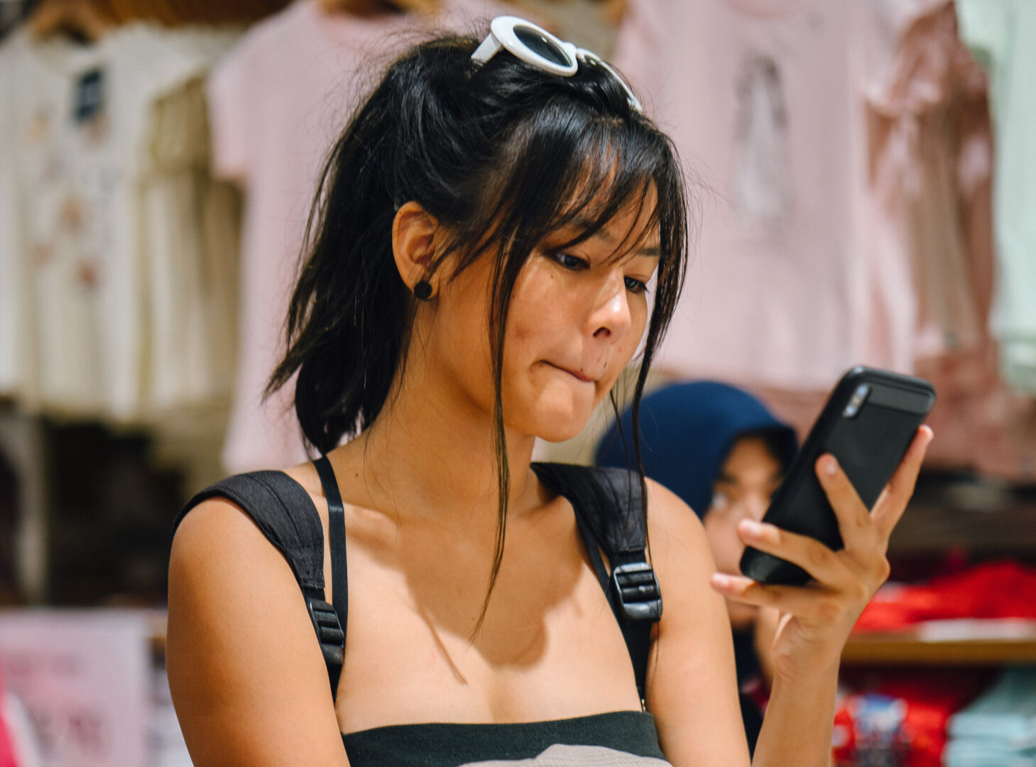 A woman with a backpack on and sunglasses on top of her head looks down on her phone. Her expression is neutral.