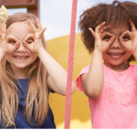 Two young girls side by side with hands over their eyes forming heart shapes.