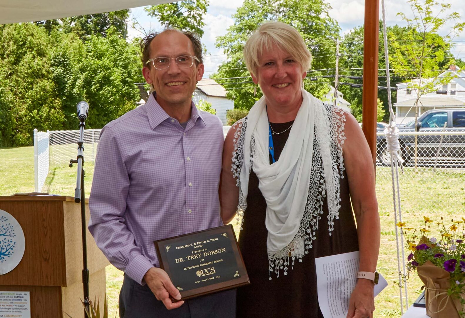 Man smiling with glasses and red shirt holding award with smiling woman with black dress.