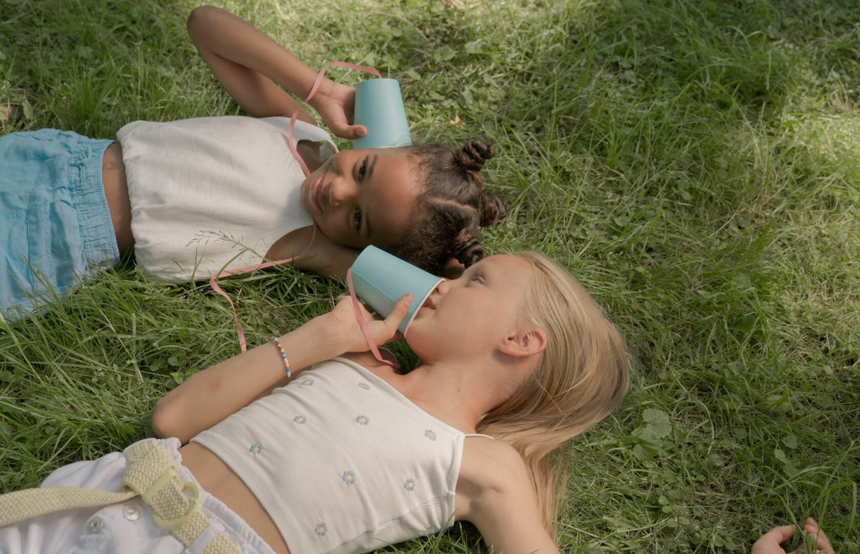 Two kids laying in the grass talking through paper cups with strings attached to them.