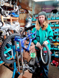 A smiling white teenage boy with long blond hair wearing a gray beanie, turquoise jacket, and blue jeans stands in a bike shop surrounded by parts bins and bicycle parts. He is holding a bicycle wheel and standing next to a blue mountain bike on a repair stand.