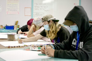 Four white teenagers, in casual attire and surgical-style face masks, sit in a row. The nearest teen is prominent in a navy hoodie. Each teen is concentrating on a drawing they are working on, on a large piece of paper on the table they are sitting at.)