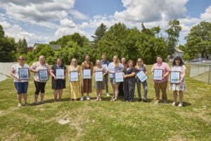 A group of UCS staff members pose for a photo while holding their Excellence Awards, against a backdrop of green grass and trees with green foliage.