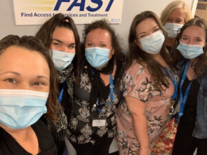 Six white women in surgical-type face masks and floral dresses stand in a row, smiling at the camera. Behind them is a small banner attached to the wall that reads, "FAST: Finding Access to Services and Treatment"
