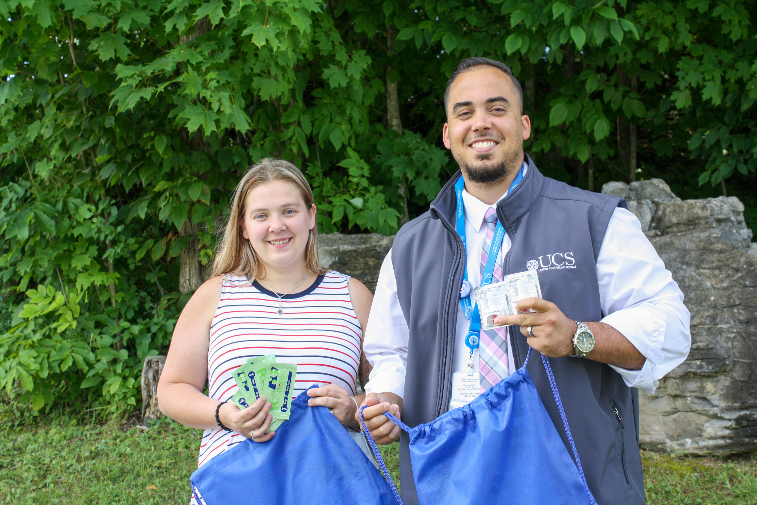 A smiling young white woman in a striped tank top and a smiling Latino man in a fleece vest stand next to each other holding blue bags and paper coupons