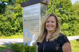 A smiling white woman with blond hair and a black short-sleeved blouse standing outdoors in front of trees, next to a large metal sign that reads “Southwestern Vermont Medical Center,” with sub-heads including a Dartmouth-Hitchcock logo and additional text reading “Centers for Living and Rehabilitation,” and “Southwestern Vermont Regional Cancer Center”)