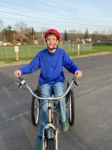 An older white teenage boy in a royal blue sweatshirt and blue jeans and wearing a red bike helmet pedals an adult-sized three-wheel cycle in an empty parking lot surrounded by a field.