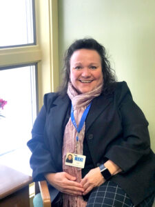 A smiling white woman with long brown hair, with a pink chiffon scarf and navy blue blazer. An ID card hangs from a royal blue ribbon around her neck