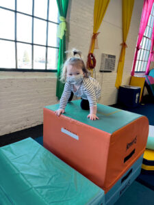 A blonde toddler girl with her hair in a top ponytail, wearing a cloth face mask and white shirt with black stripes and black leggings, looks at the camera while crawling on top of a large foam platform block in front of a white-painted brick wall with a window and wall air-conditioner.