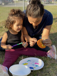A closeup of a curly brown-haired, Brown toddler girl in a black tank top and maroon leggings sitting on a lawn holding a paintbrush, straddling a paper plate of paint on the grass in front of her; kneeling beside her is a Brown woman with her brown hair in a topknot, holding a fist-sized pumpkin she is helping the toddler to paint.