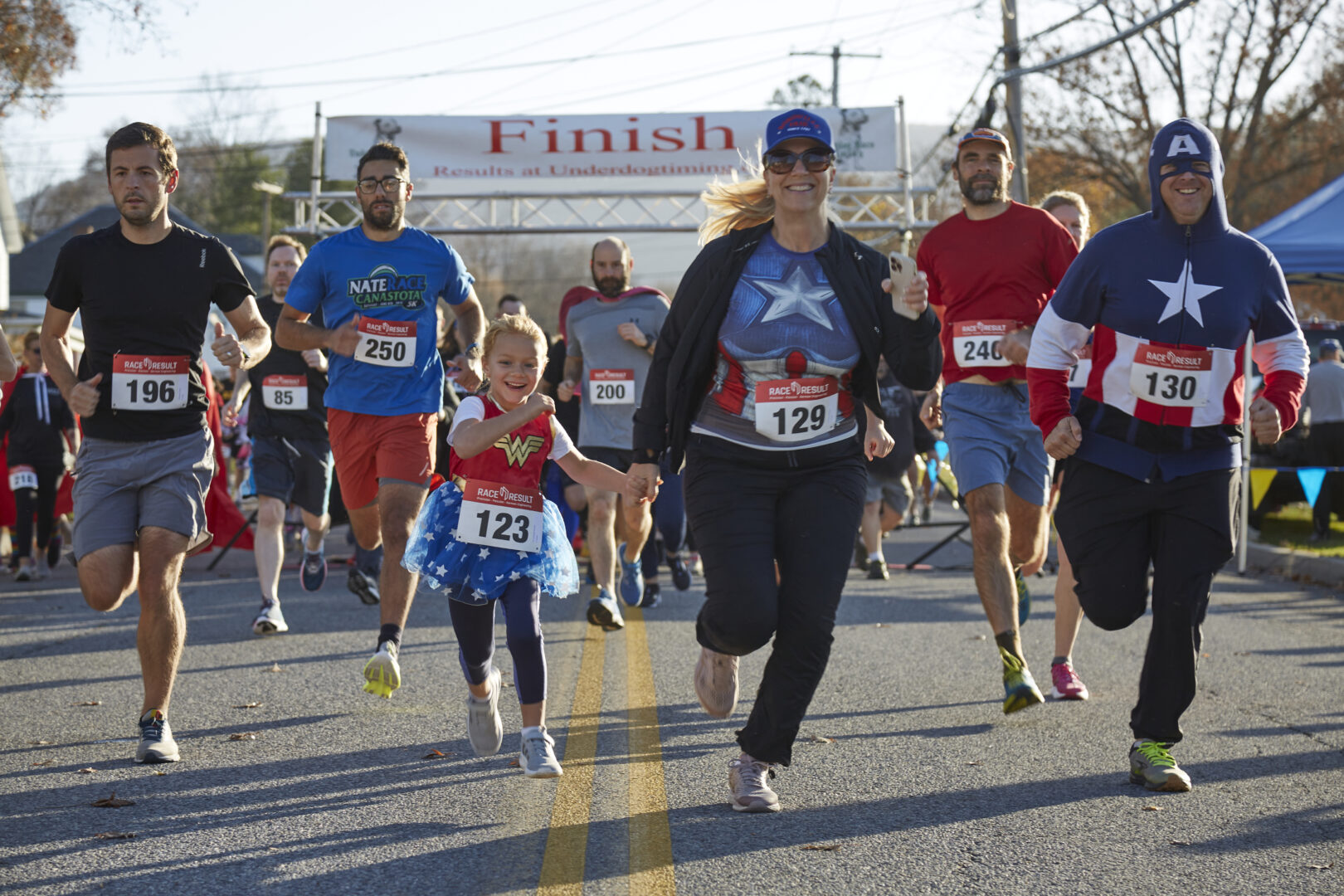 Little girl dressed in Supehreo Costume holding woman in superhero costume's hand at start of race. Other racers pictured in background.