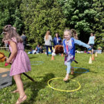 A young child in a pink dress and a young child in blue pants and a blue shirt jump in hula hoops on the grass.