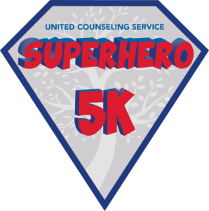 Logo in the shape of a diamond outlined in navy blue. Inside the diamond in all caps small text is “UNITED COUNSELING SERVICE” in navy blue above large 3-D all-caps cartoon-style lettering in red with navy dimensions, “SUPERHERO 5K”. The text is set over a graphic of a pale gray stylized tree knocked out of the mid-tone gray background. Across the bottom of the diamond is a rectangular banner with a navy blue border, with the words “5th Annual” in white on a red background.