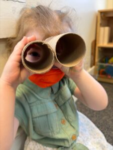 A closeup of a blonde toddler in a green dress and red face mask holding up a pair of “binoculars” made up of two paper-towel holders taped together