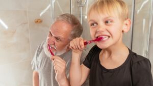 Closeup of a young white boy with blond hair in a black t-shirt brushing his teeth with a red plastic toothbrush; to his left leans an older white man with short gray hair and a short white beard in a gray t-shirt, also brushing his teeth with a red plastic toothbrush.