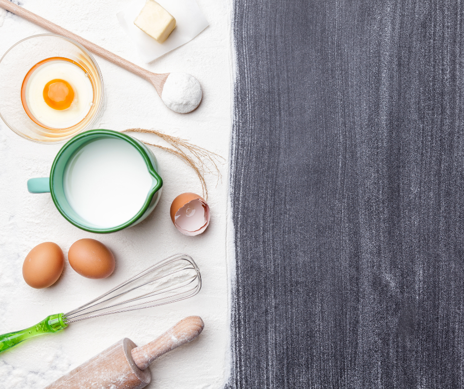 Wooden spoons, a whisk, a pitcher of milk, whole eggs, and cracked eggs rest on a white background, with half the photo a simple gray countertop background.