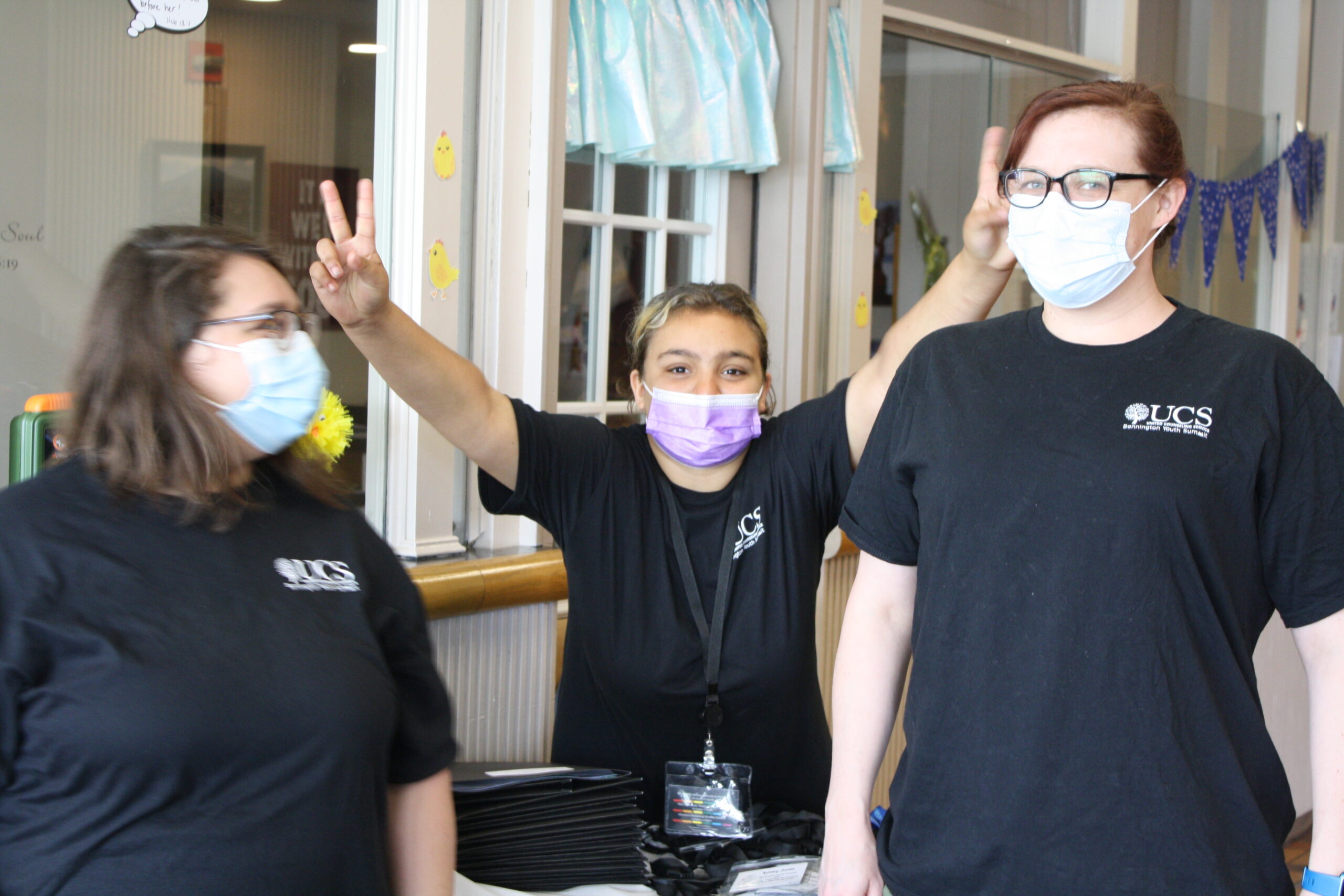 Young person with brown hair and blonde highlights makes peace symbols with hands while standing between two people. All three people are wearing black t-shirts that ready, "UCS Bennington Youth Summit."