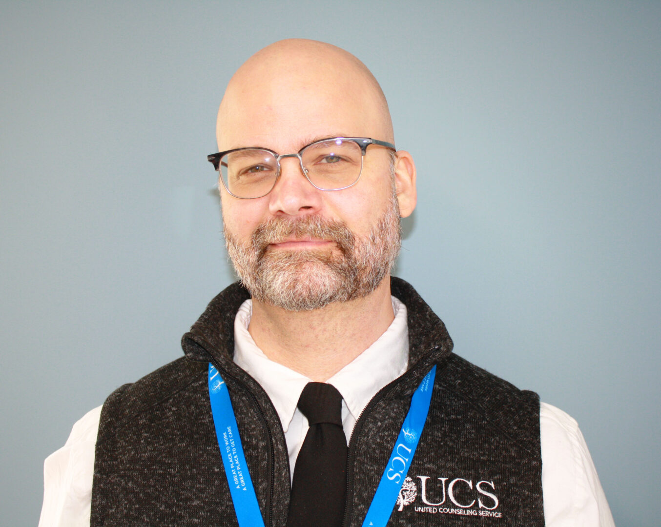 A man with glasses wearing a black UCS vest, black tie, blue lanyard, and white shirt smiles.