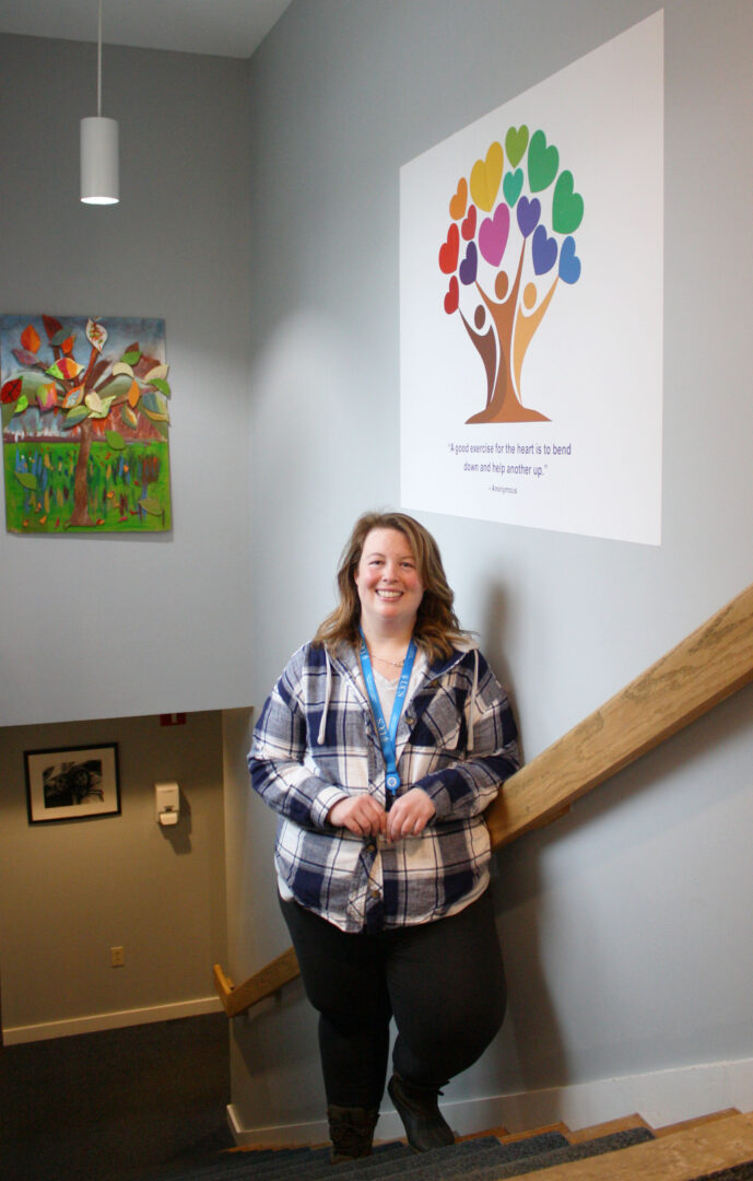 A smiling woman stands on a staircase near two pieces of art featuring colorful tree graphics.