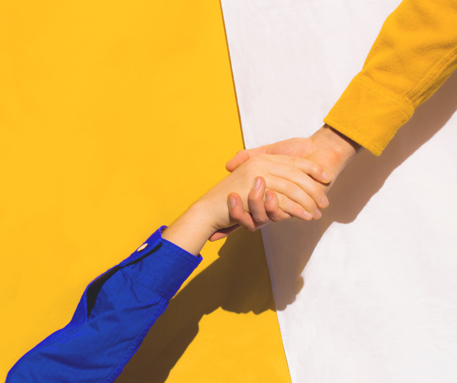 an arm in a blue sleeve reaches and grabs a hand with a yellow-sleeved arm against a split yellow and white background.