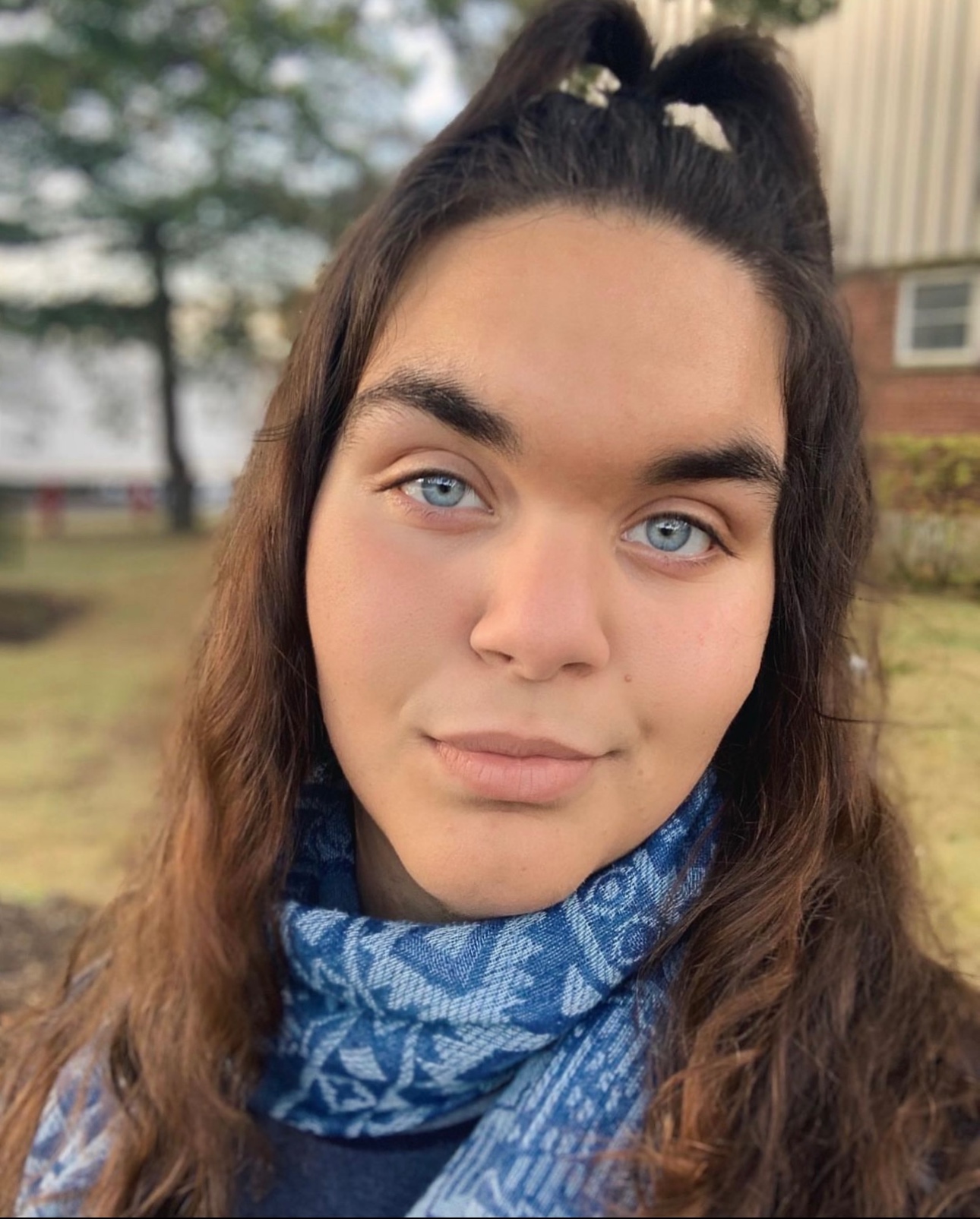 A person with long brunette hair, blue eyes, and a blue patterned scarf against a background of green grass and a brick and tan building