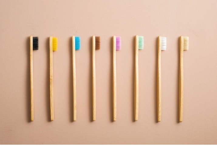 Eight wooden toothbrushes lined up against a peach colored background. Each toothbrush has a different color bristles.