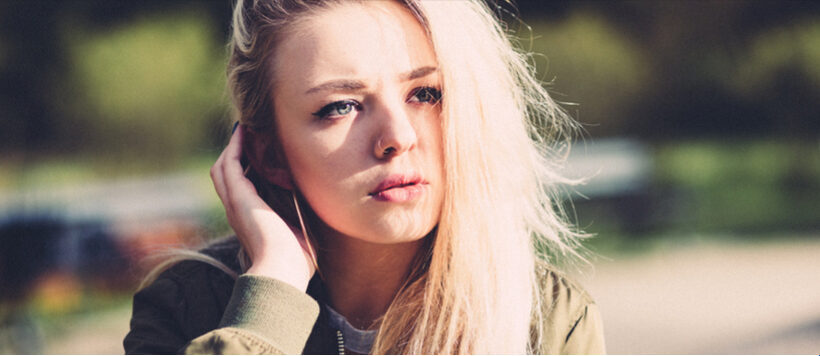 a young woman with long blonde hair, blue eyes, and earbuds, looking into the distance