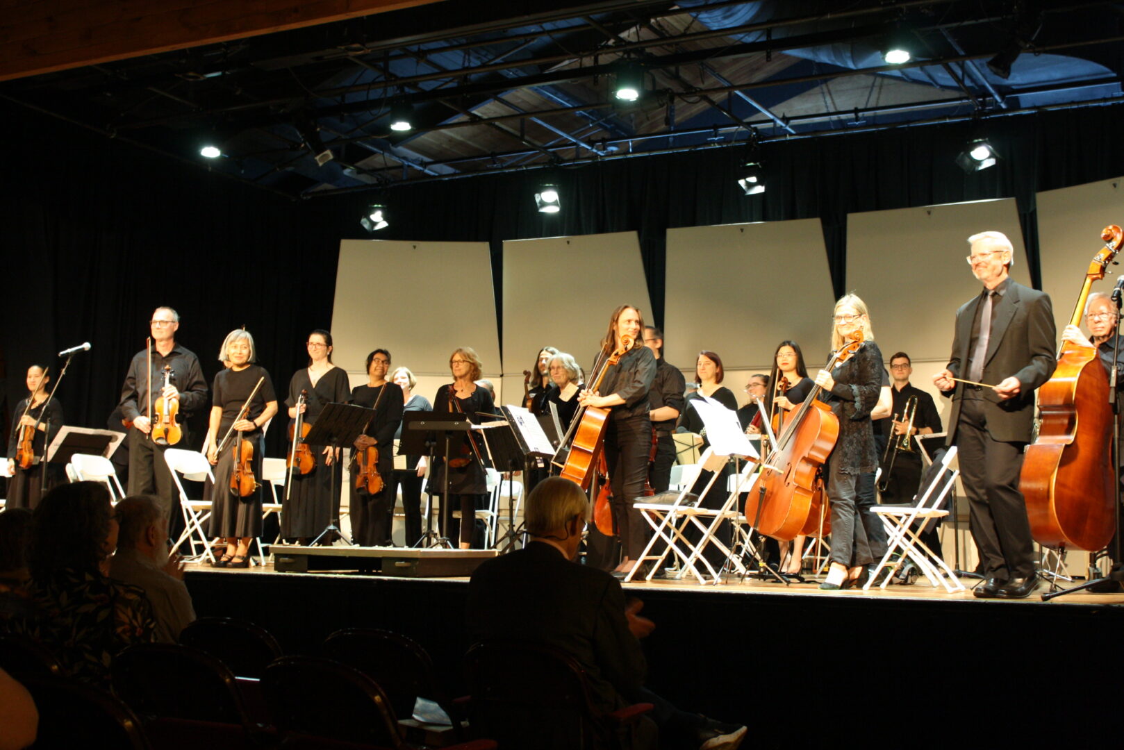 An orchestra stands facing an audience. The front row of the orchestra features violinists and cellists.