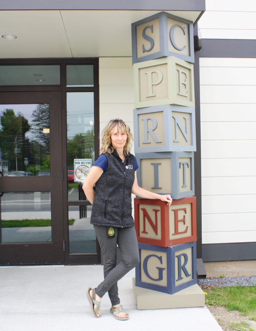 A woman with long blonde hair wearing a dark gray vest with a UCS logo on it stands against a building with a column made out of large building blocks that spell SPRING.