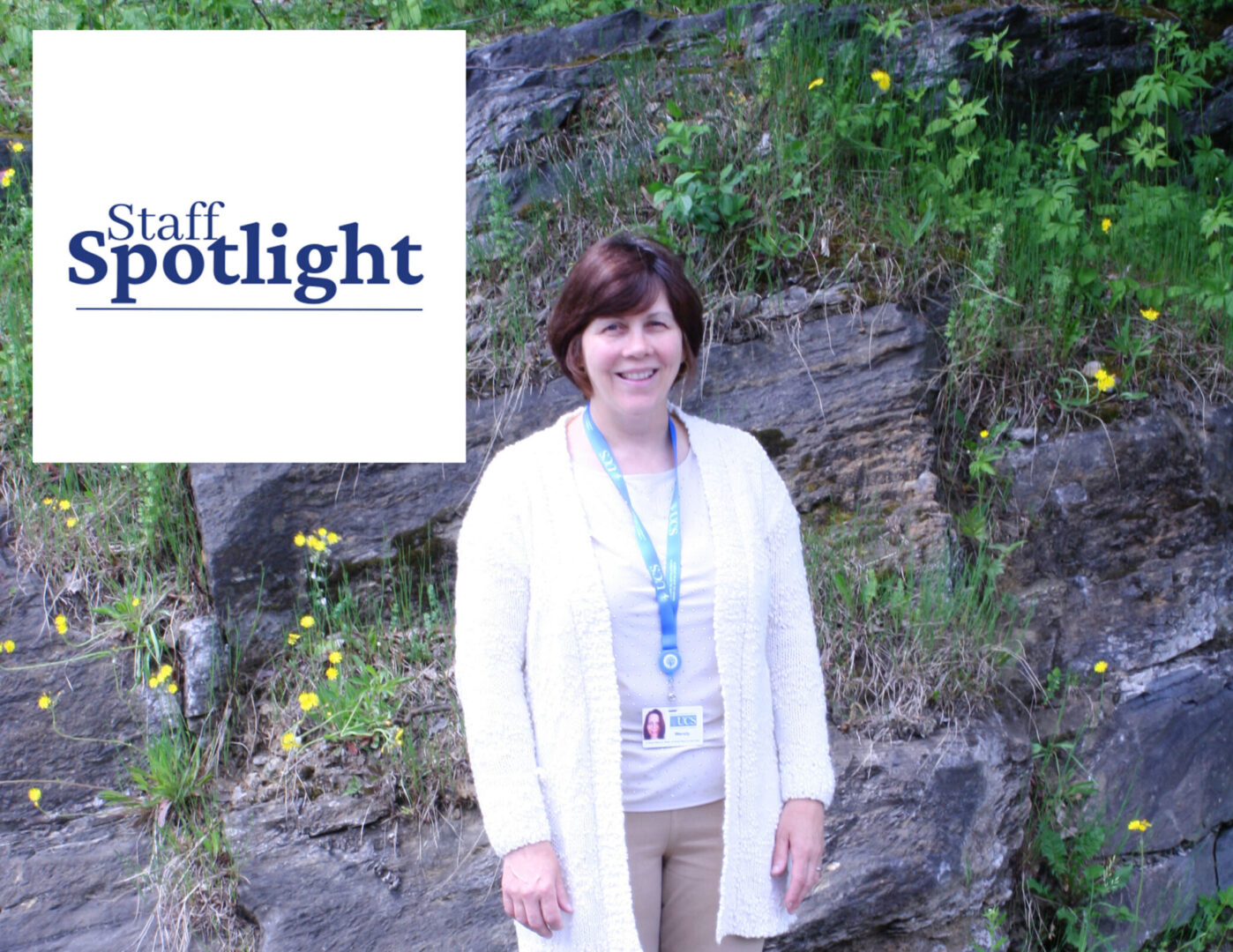 A woman with short brunette hair wearing a white shirt and sweater and tan pants stands against a rocky background, A graphic with "Staff Spotlight" is in the top left.