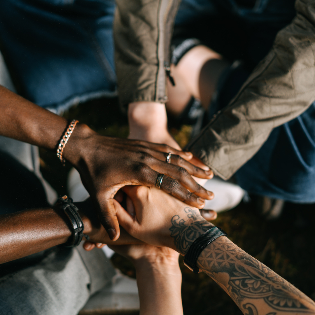 several hands on top of each other. One arm has tattoos, and one has rings and a bracelet. One has a black watch.