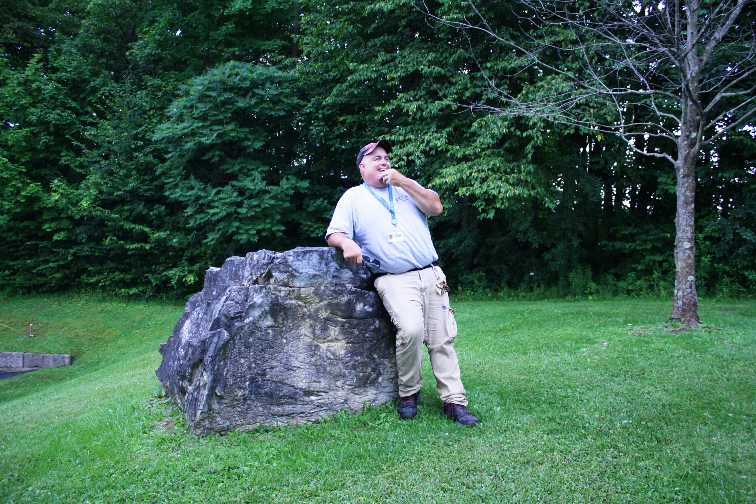 A smiling man with his left hand up to his chin leans against a boulder. Background has green grass and trees, and a small maple tree.