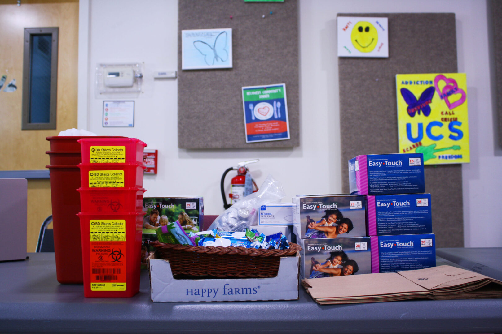 Harm reduction and syringe services supplies sit on a table. These include red sharps disposal containers, clean syringes in blue boxes, alcohol prep pads, and rice krispies treats.