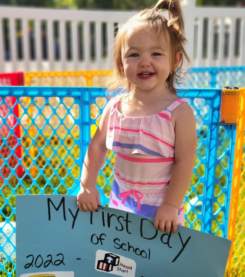 A pre-school age little girl in a pink striped outfit and a ponytail smiles while holding a blue sign that says My First Day of School 2022-2023.