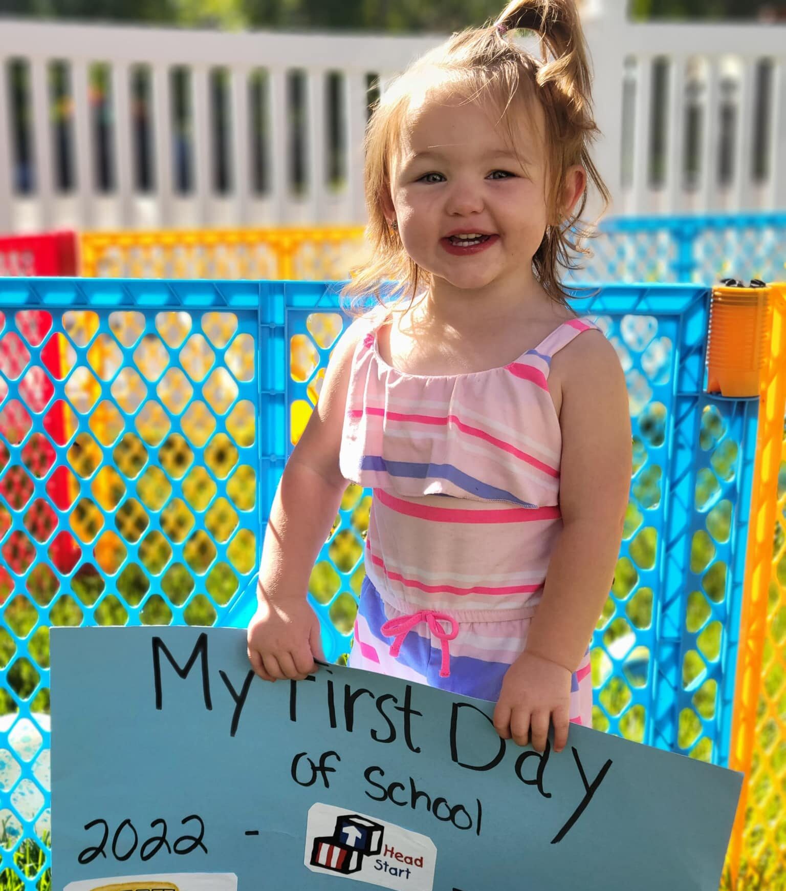 A pre-school age little girl in a pink striped outfit and a ponytail smiles while holding a blue sign that says My First Day of School 2022-2023.