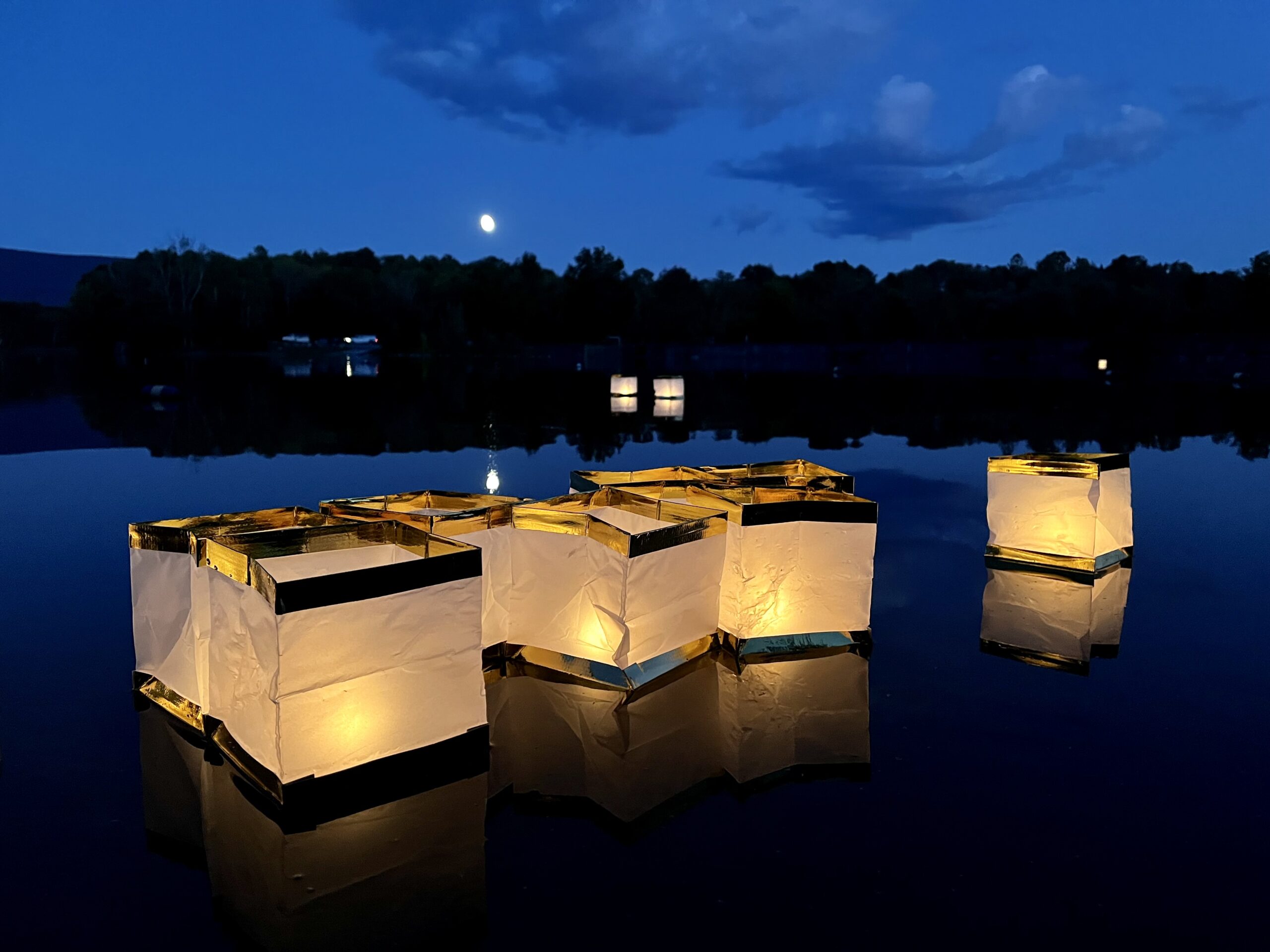 Candle-lit paper lanterns float on a dark lake against a darkening blue sky with the outline of a treeline and a full moon in the background.