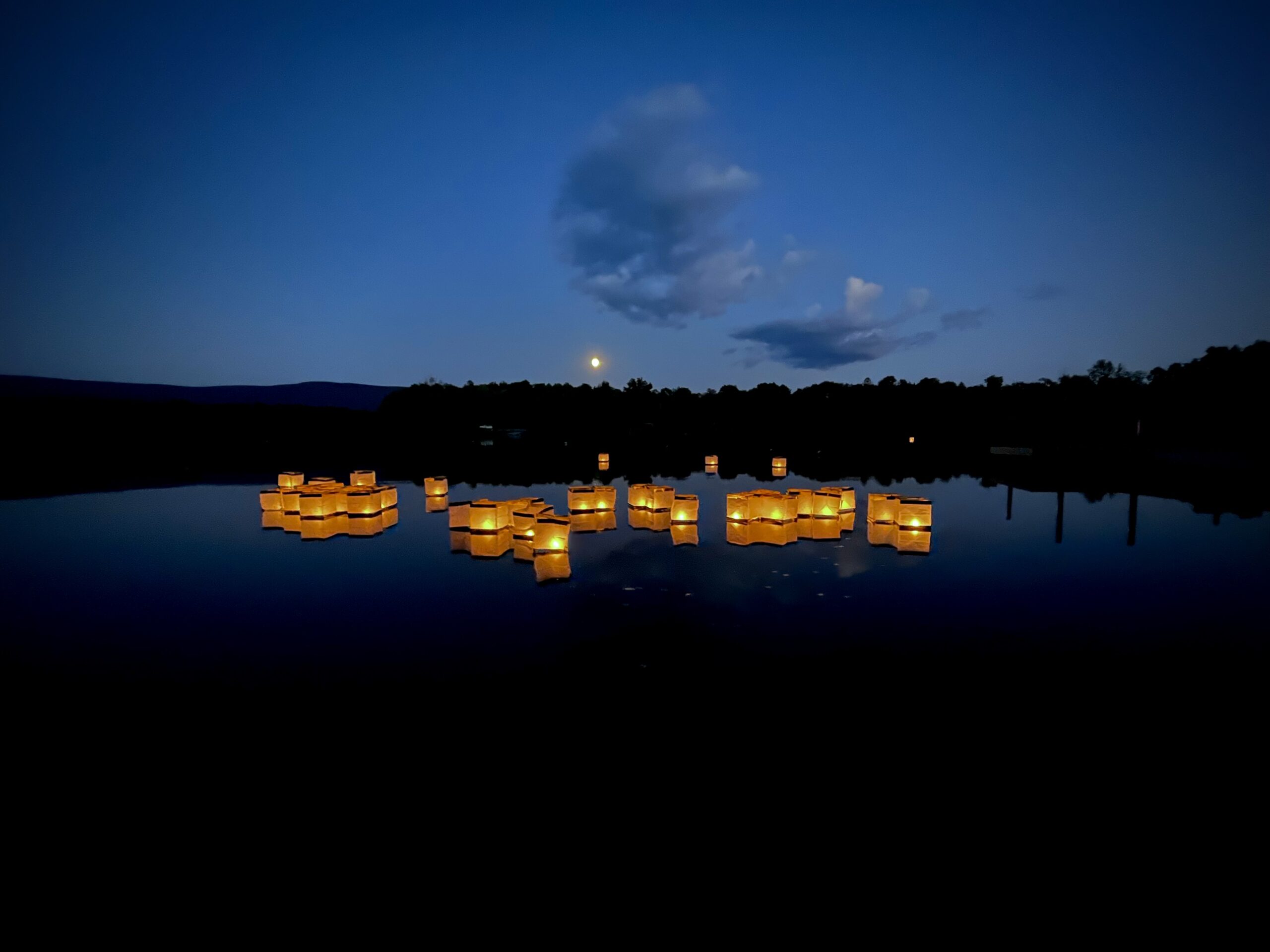Cube shaped paper lanterns lit with candles float on a dark lake against a darkening blue sky. There is a bright full moon in the background and a couple light clouds. A tree line is also visible just below the moon.