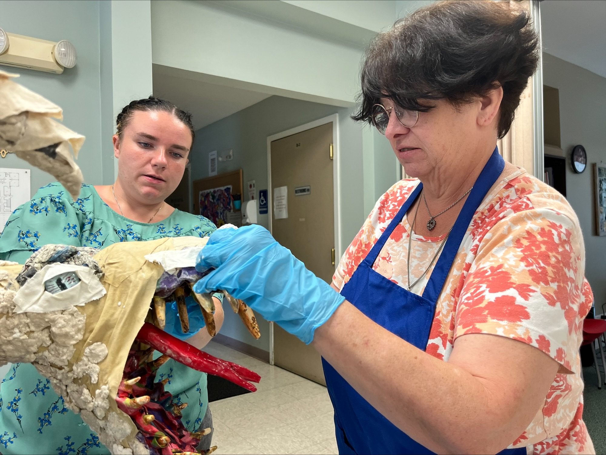 A woman with short dark hair works on a dragon art project. A young woman looks on.