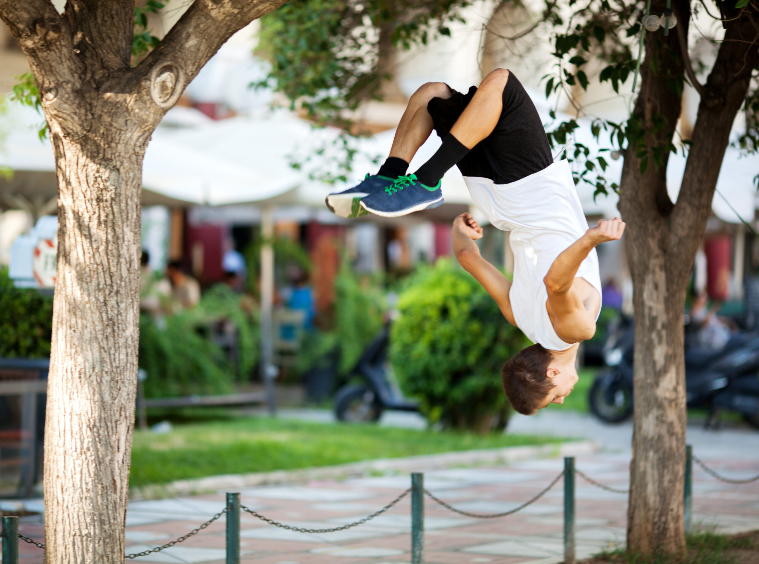 A young man wearing a white t shirt and black shorts and blue sneakers foes a front flip against an urban park background.